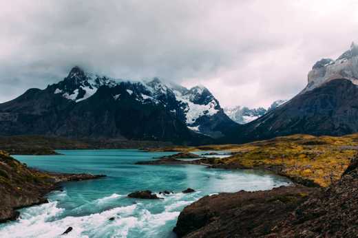 Turquoise glacial waters reflect rugged, snow-capped peaks beneath a moody sky in Torres del Paine National Park, Chile. 