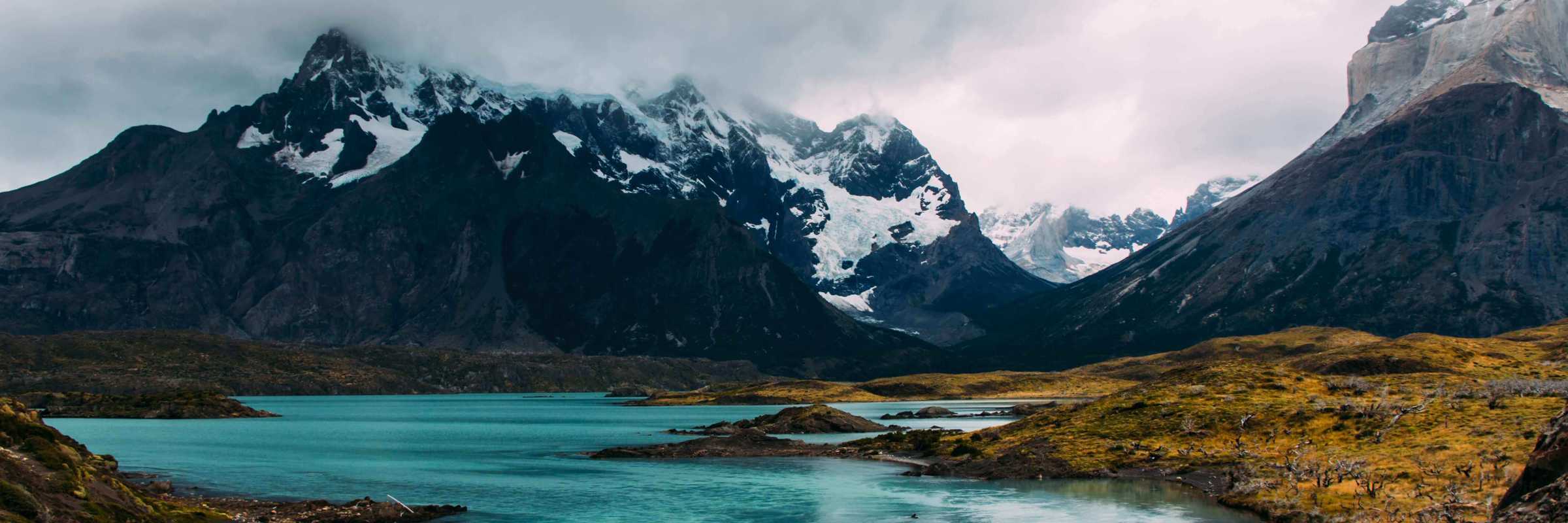Turquoise glacial waters reflect rugged, snow-capped peaks beneath a moody sky in Torres del Paine National Park, Chile. 