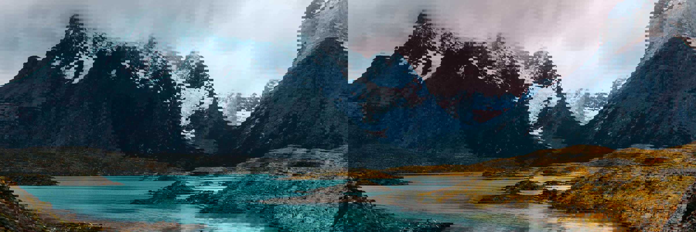 Turquoise glacial waters reflect rugged, snow-capped peaks beneath a moody sky in Torres del Paine National Park, Chile. 