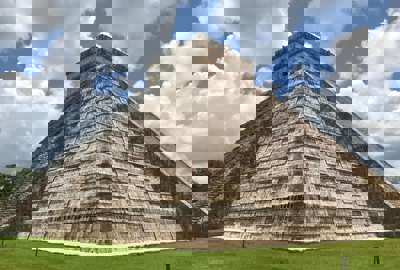 Temple of Kukulcan pyramid at Chichen Itza, Yucatan, Mexico, with cloudy blue sky and green grass foreground. 