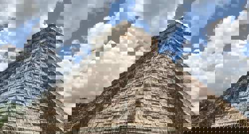 Temple of Kukulcan pyramid at Chichen Itza, Yucatan, Mexico, with cloudy blue sky and green grass foreground. 
