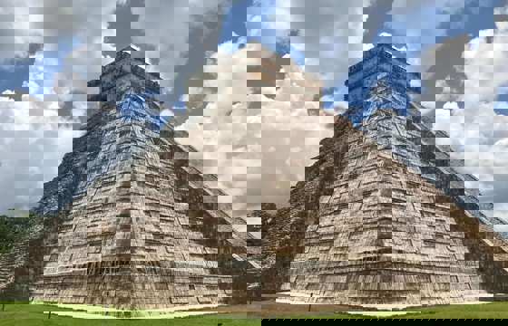 Temple of Kukulcan pyramid at Chichen Itza, Yucatan, Mexico, with cloudy blue sky and green grass foreground. 