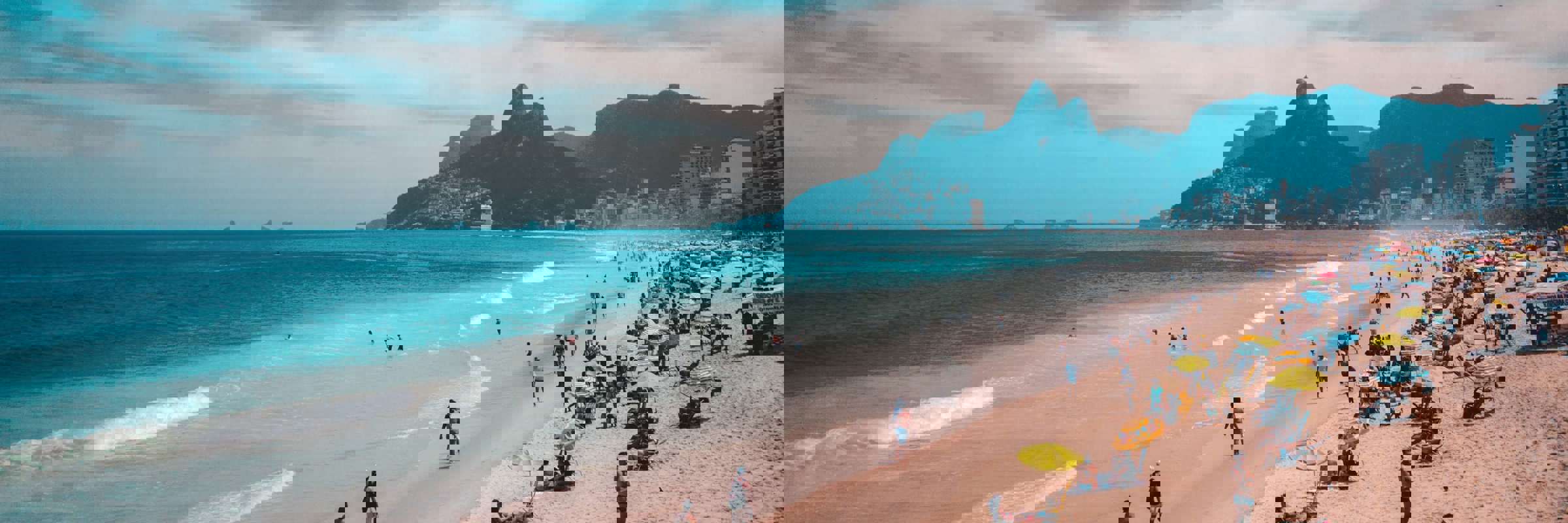 View of Ipanema Beach with beachgoers, ocean waves, and the Two Brothers Mountain in Rio de Janeiro, Brazil. 