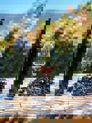 A bottle of red wine, a glass, and a cluster of grapes sit on a vineyard table with Chilean vines and hills in the background. 