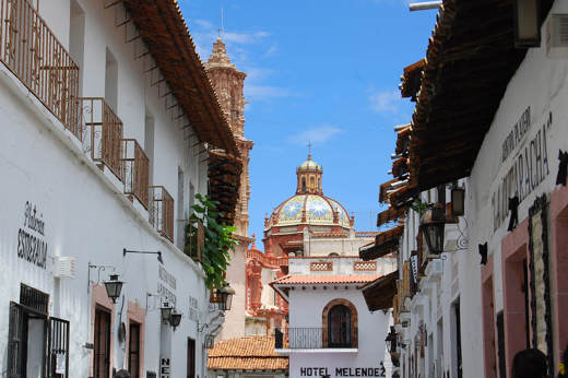 Whitewashed colonial street with balconies, lanterns, and a distant church dome in Taxco, Mexico. 