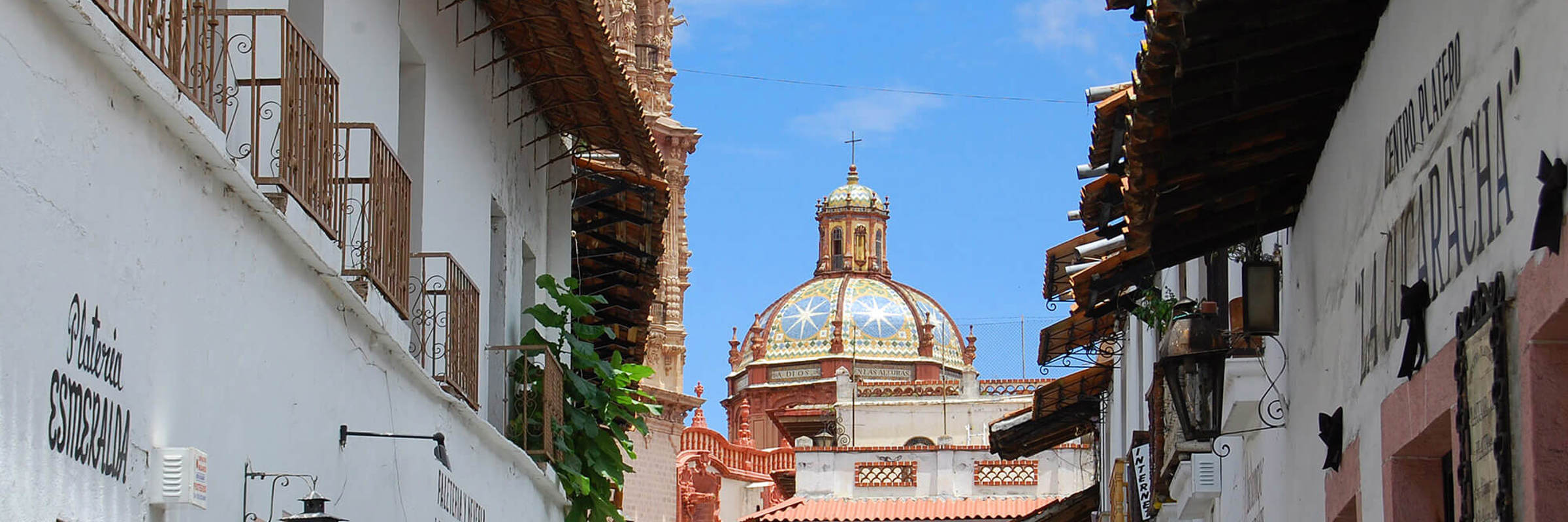 Whitewashed colonial street with balconies, lanterns, and a distant church dome in Taxco, Mexico. 