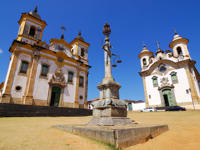 Twin colonial churches and monument in Tiradentes, a historic town in Minas Gerais, Brazil, under a clear blue sky. 