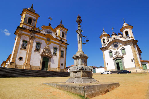 Twin colonial churches and monument in Tiradentes, a historic town in Minas Gerais, Brazil, under a clear blue sky. 