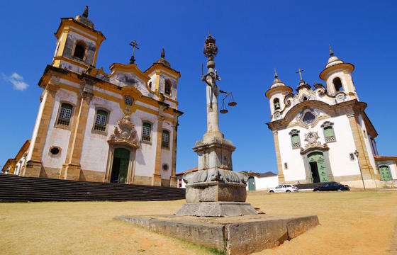 Twin colonial churches and monument in Tiradentes, a historic town in Minas Gerais, Brazil, under a clear blue sky. 