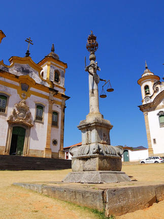 Twin colonial churches and monument in Tiradentes, a historic town in Minas Gerais, Brazil, under a clear blue sky. 