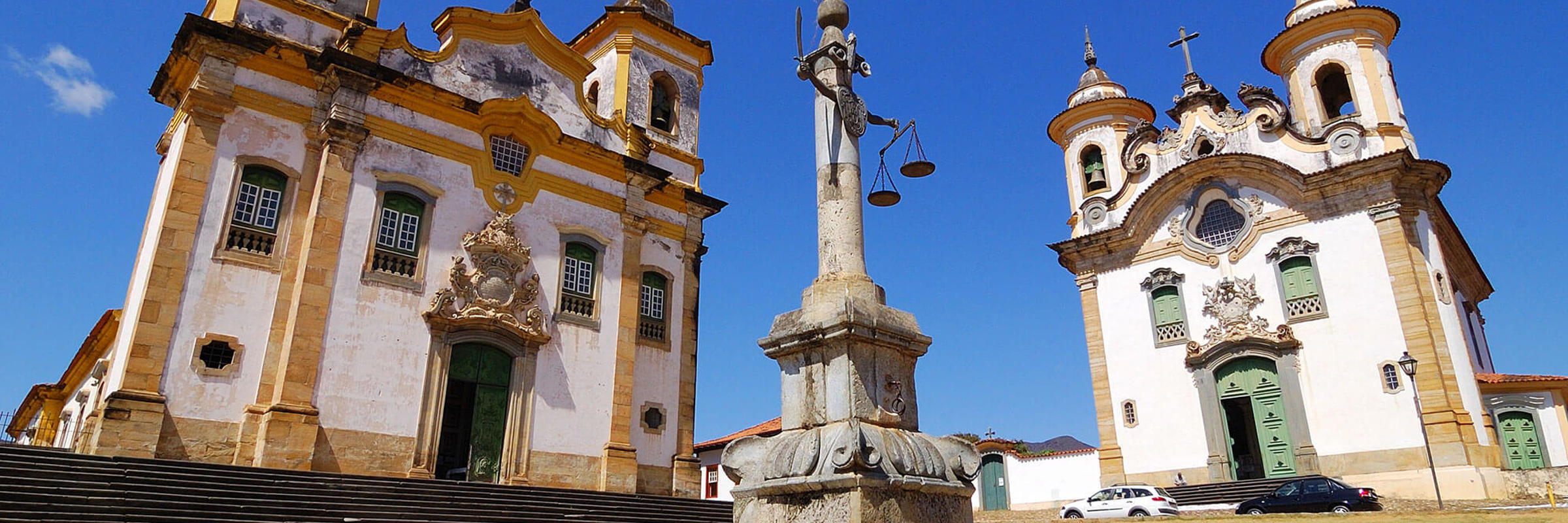 Twin colonial churches and monument in Tiradentes, a historic town in Minas Gerais, Brazil, under a clear blue sky. 