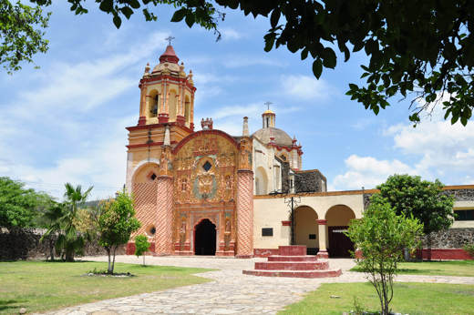 Colonial red-brick church in Queretaro, Mexico, surrounded by trees and blue sky, showcasing local architecture. 