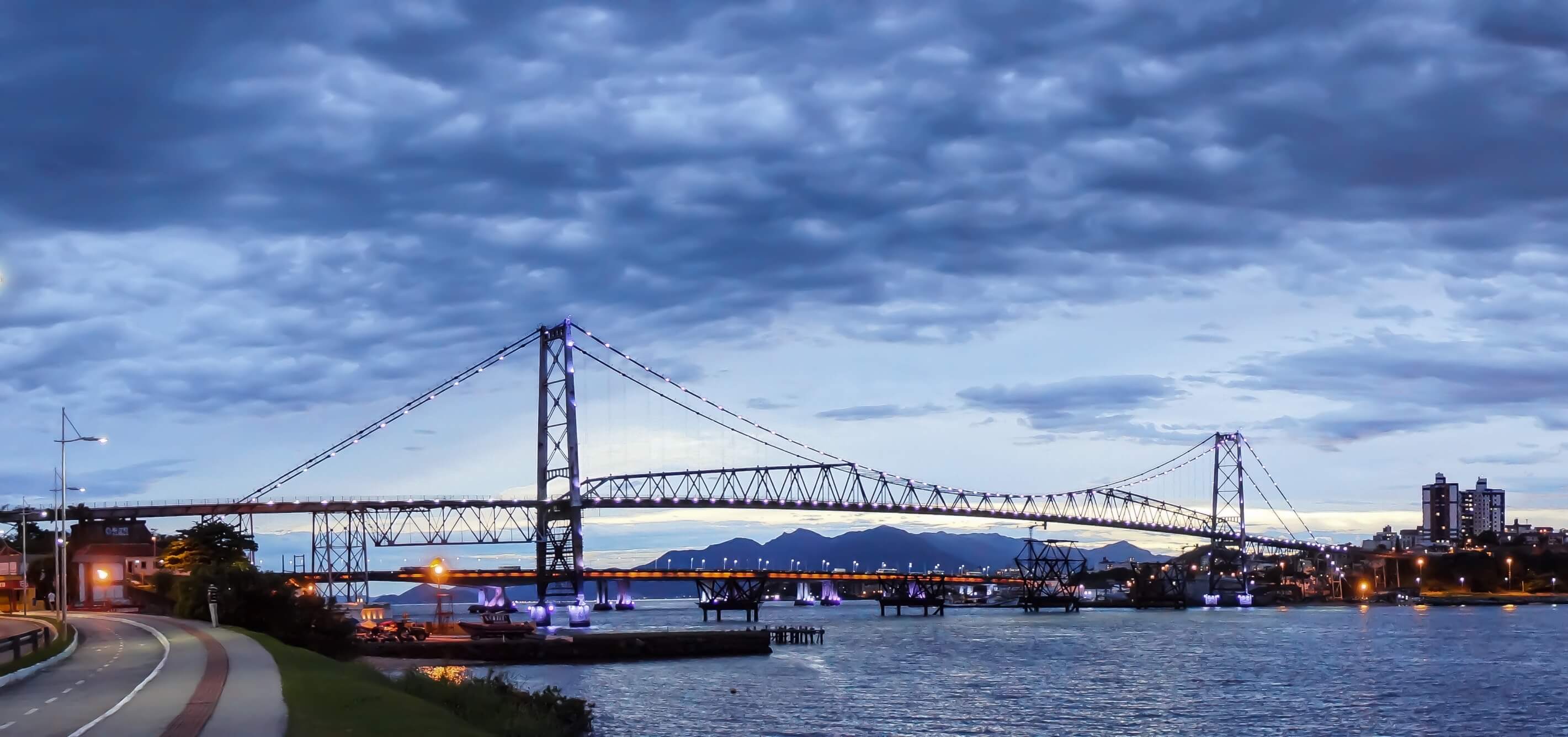 The Hercilio Luz Bridge at sunet in Florianopolis, Brazil, spanning the water with city lights and dramatic evening sky. 