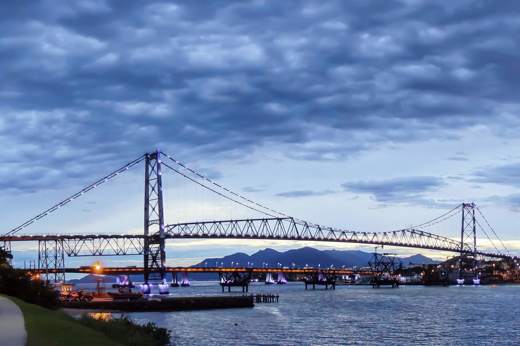 The Hercilio Luz Bridge at sunet in Florianopolis, Brazil, spanning the water with city lights and dramatic evening sky. 