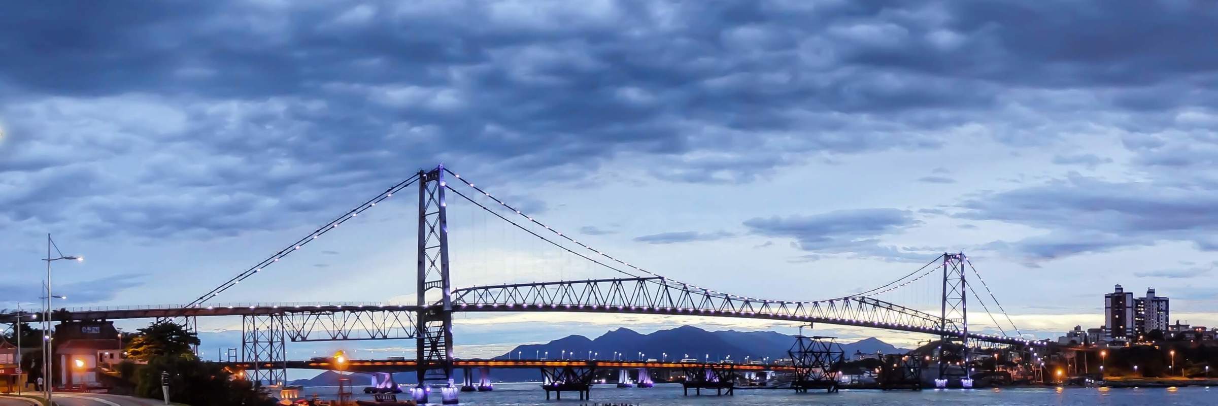 The Hercilio Luz Bridge at sunet in Florianopolis, Brazil, spanning the water with city lights and dramatic evening sky. 