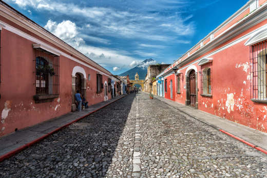 Cobblestone street in Antigua, Guatemala with pastel houses and a distant volcano under blue skies. 