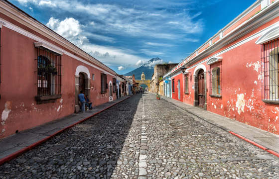 Cobblestone street in Antigua, Guatemala with pastel houses and a distant volcano under blue skies. 
