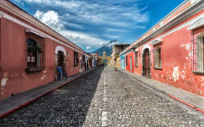 Cobblestone street in Antigua, Guatemala with pastel houses and a distant volcano under blue skies. 