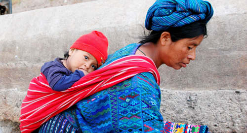Central America - Guatemala, Indigenous mother carries child in traditional woven clothing, reflecting daily life and culture. 