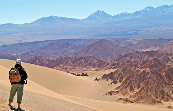 Hiker explores the vast, sunlit dunes of Chile's Atacama Desert with Andes backdrop. 
