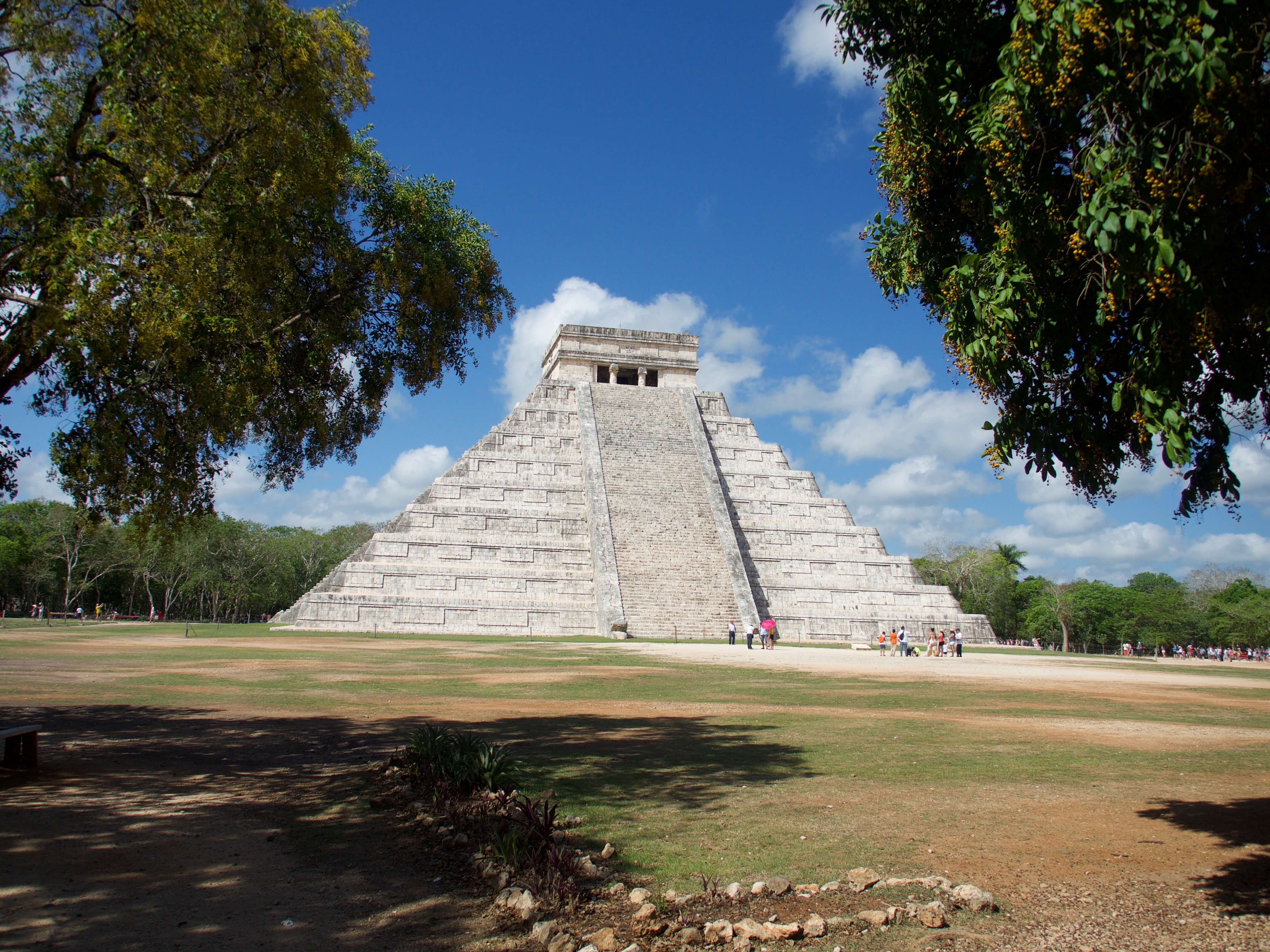 The Temple of Kukulkan, also known as El Castillo, stands tall at Chichen Itza under a bright blue Yucatan sky surrounded by trees. 