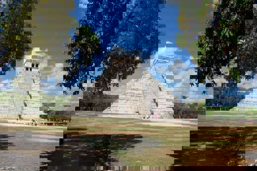 The Temple of Kukulkan, also known as El Castillo, stands tall at Chichen Itza under a bright blue Yucatan sky surrounded by trees. 