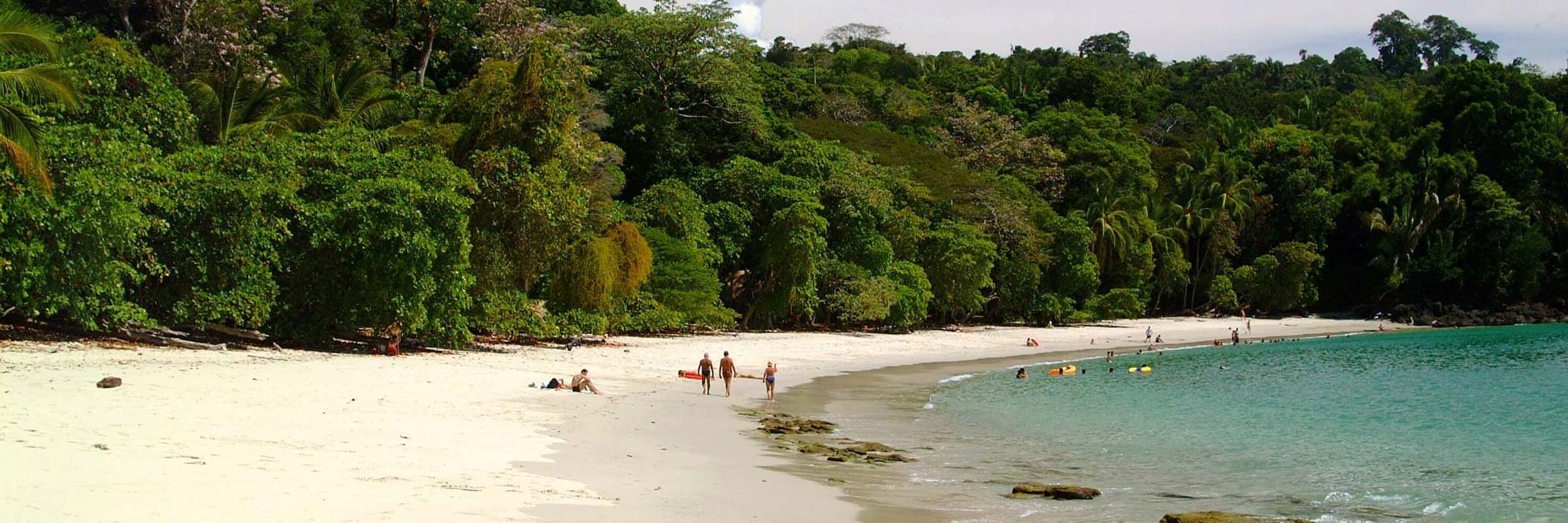 White sandy beach backed by lush rainforest in Manuel Antonio National Park, with people walking along the the shore and calm turquoise waters. 