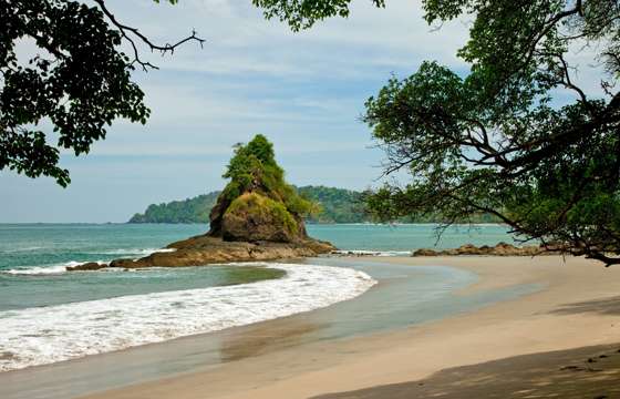 A rocky islet rises just offshore as gentle waves lap the peaceful sands of Manuel Antonio. 