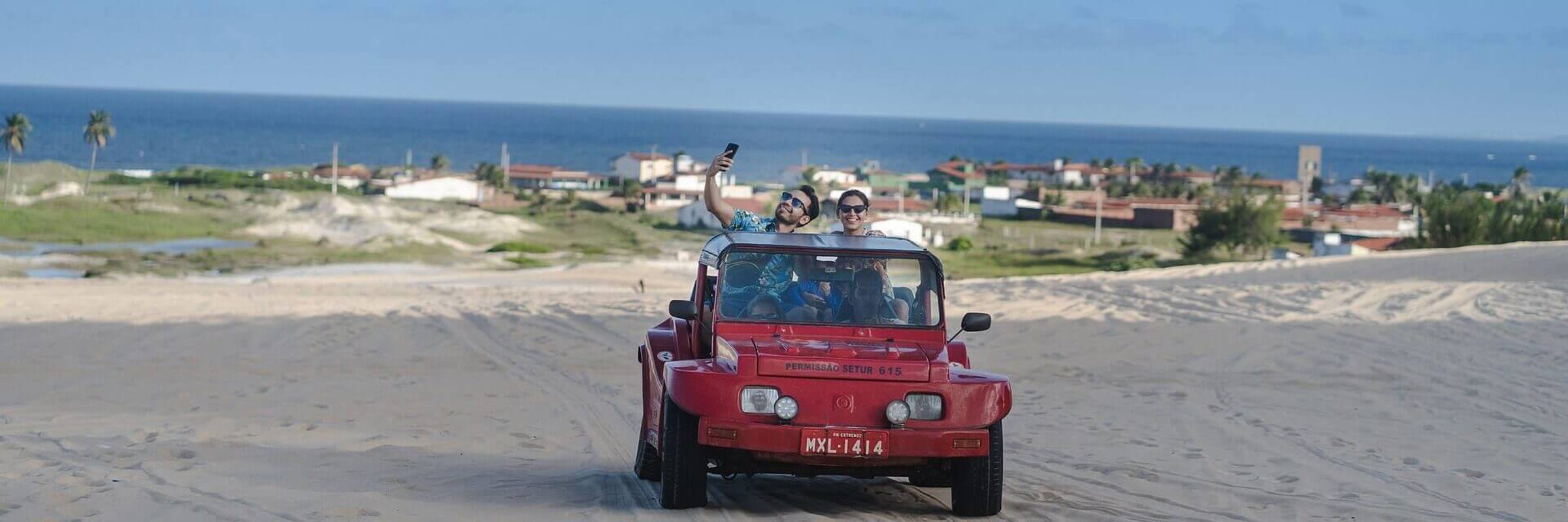 Red beach buggy driving  across sand dunes in Jericoacoara with ocean and coastal town in the background. 